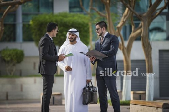 Middle Eastern man carrying a laptop bag talking to his colleagues|-
