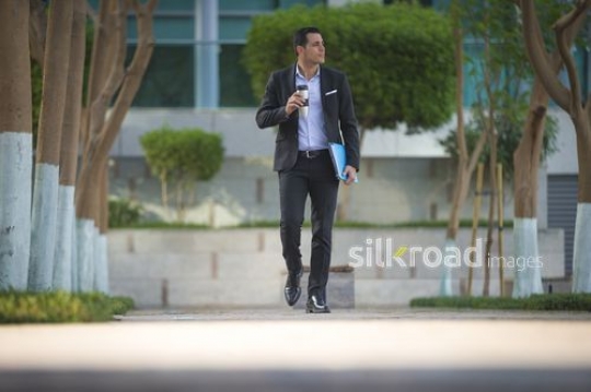 Middle Eastern man walking outside carrying a coffee mug and folders|-