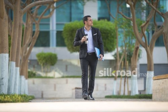 Middle Eastern Businessman walking outside carrying a travel mug and folders|-