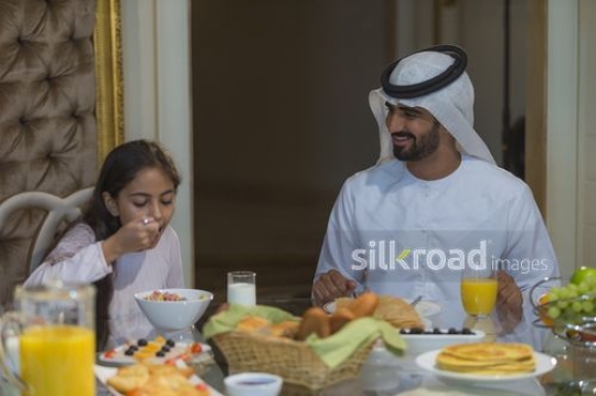 Young arab girl eating breakfast sitting next to her father at the dining table|