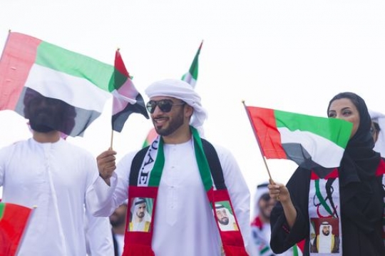 Men and woman dressed in the traditional UAE attire carrying a UAE Flag|