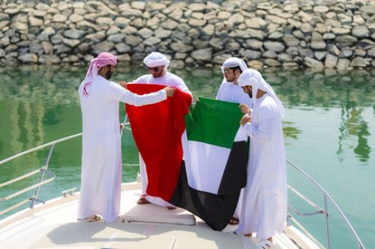 Arabian men standing on the deck of the yacht holding the UAE flag to celebrate National Day|