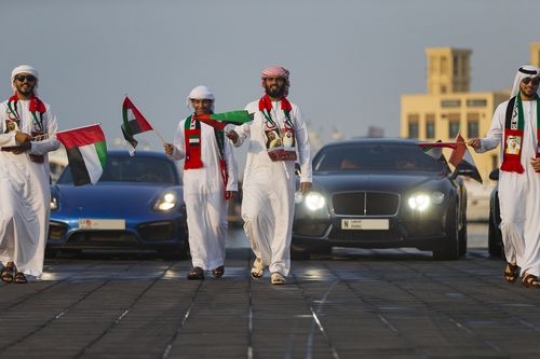 A group of men dressed in the UAE traditional attire followed by luxury vehicles during a National Day parade|