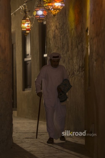 Arab man carrying a prayer rug and walking through the alleyway of Ramadan Lanterns|-