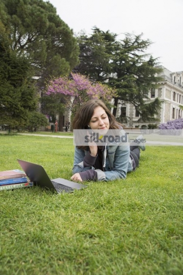 Woman laying down on the grass using the laptop talking on the phone|