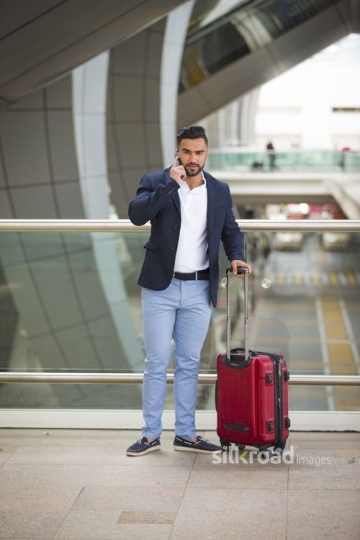Middle Eastern man standing with his luggage using his mobile|-